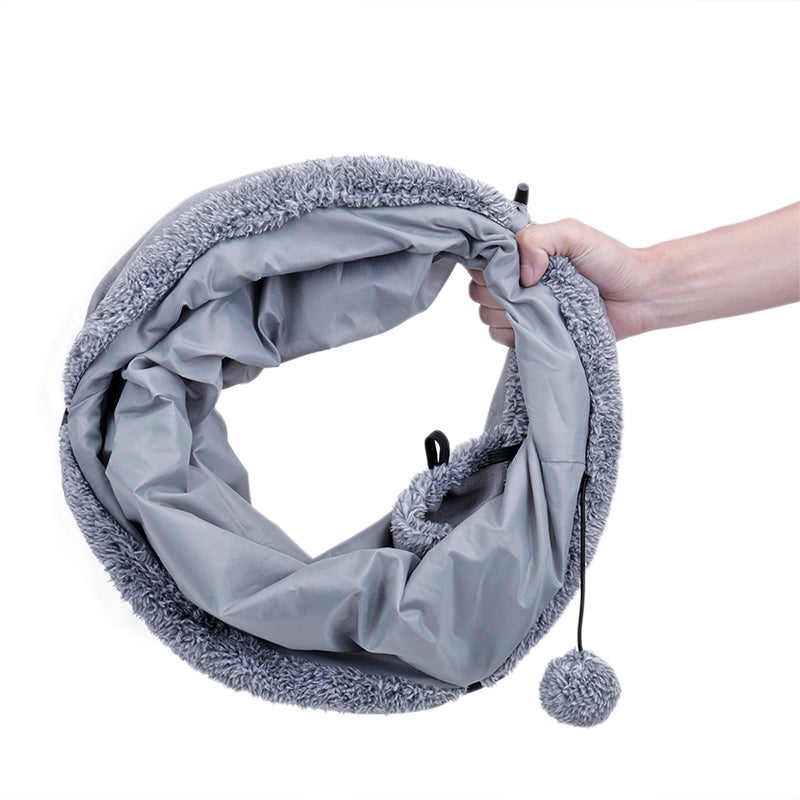 Hand holding the grey suede-touch cat tunnel folded flat for storage, with a pom-pom toy