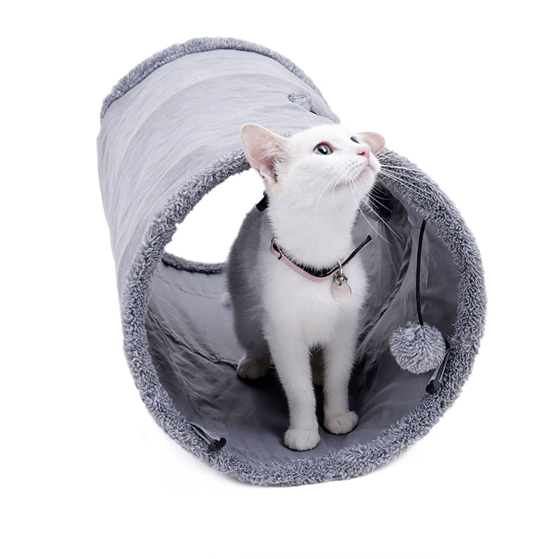 White cat standing in a grey suede-touch cat tunnel looking up at a pom-pom toy