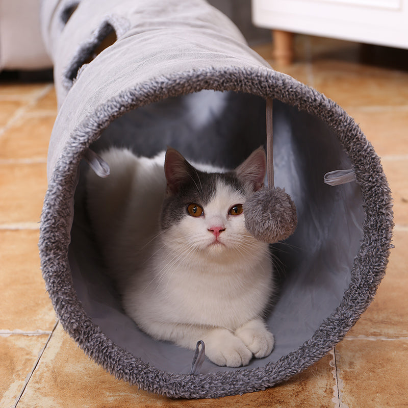 Kitten looking out from a grey suede-touch cat tunnel with a pom-pom toy